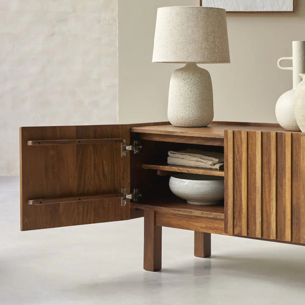 Wooden sideboard with open door displaying interior, lamp, and decorative items on a neutral background.