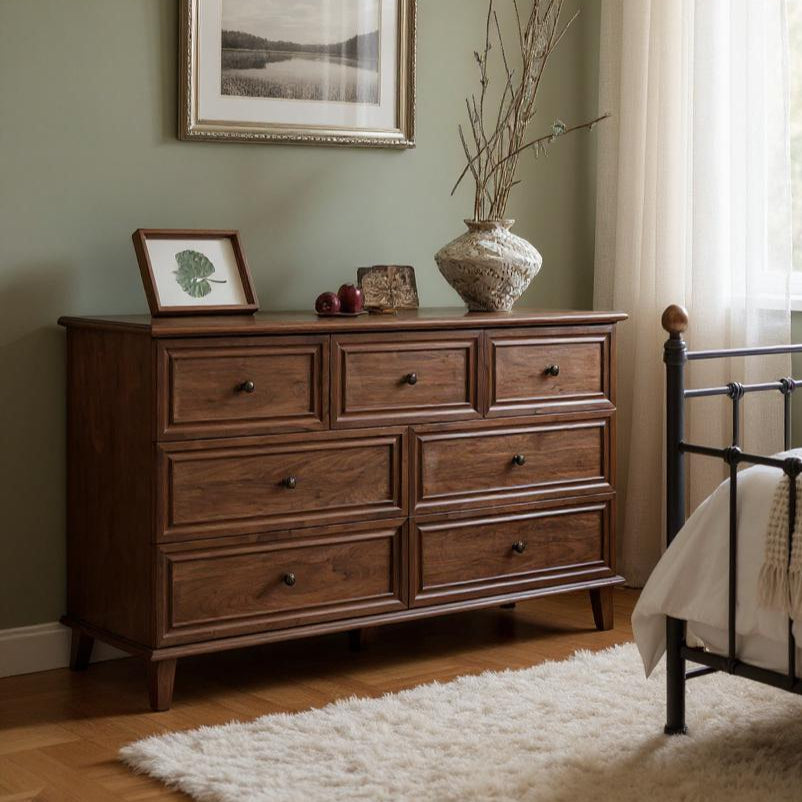 Wooden dresser in a bedroom with a bed and framed picture on the wall.